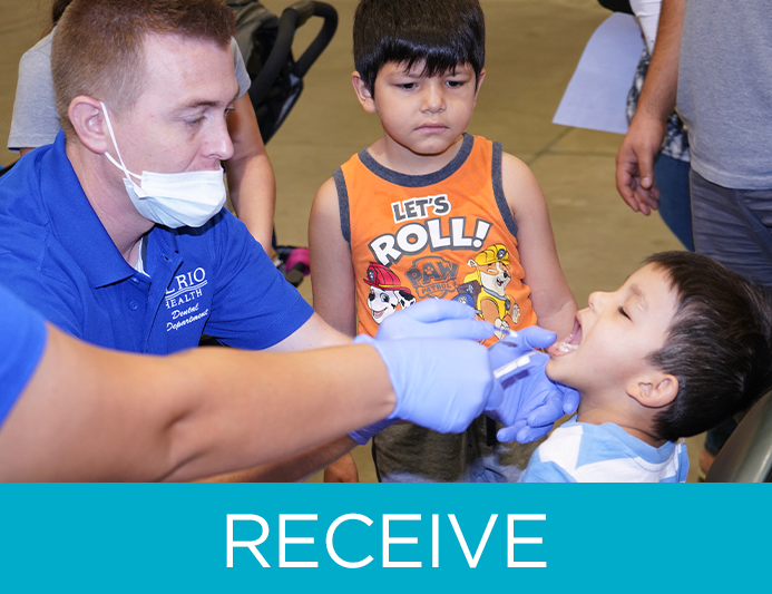 two oral health professionals look inside a young boy's mouth as another young boy looks on during an oral health volunteer event 